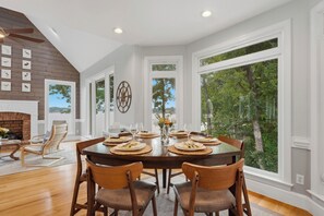 A dining room with a large table set for six, surrounded by wooden chairs, large windows with a view of trees, and a fireplace with a brick surround. - Lakefront + Private Dock | Hot Tub + Pool Table + Cabana | Point of View (Mooresville)