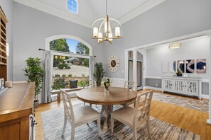 Een eetgedeelte met een ronde tafel, stoelen, een kroonluchter en uitzicht op een huis door een groot raam.