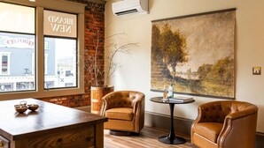 Une pièce avec une table en bois, deux fauteuils en cuir, une table ronde avec une bouteille et des verres, une grande peinture abstraite et une vue sur un bâtiment par la fenêtre.