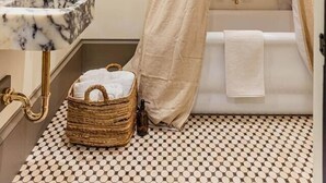 Une salle de bain avec un lavabo en marbre, un robinet doré, une baignoire avec un rideau beige, un petit panier en osier et un carrelage à motifs.