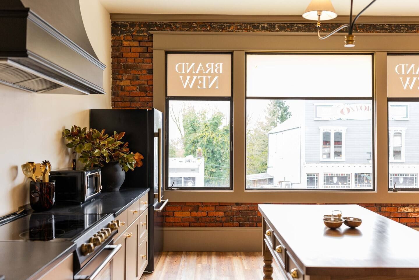 A kitchen with a brick wall, a large window, and a countertop with a plant and kitchenware.