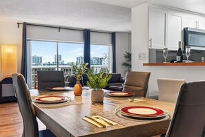 A modern dining area with a wooden table set for four, surrounded by chairs, with a city view through large windows. - Sunny Isles Condo • Shared Pool + Private Balcony • Near Beaches | Isle Vista (Sunny Isles Beach)