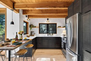 A modern kitchen with dark cabinetry, a stainless steel refrigerator, and a wooden ceiling. - Cannon Beach Stay + Treetop Deck + Walk To Beach + Haystack Rock | Salt & Spruce (Cannon Beach)