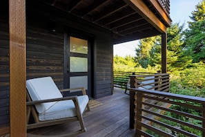 A wooden deck with two white cushioned chairs, a wooden railing, and a door leading to a covered porch. - Cannon Beach Stay + Treetop Deck + Walk To Beach + Haystack Rock | Salt & Spruce (Cannon Beach)