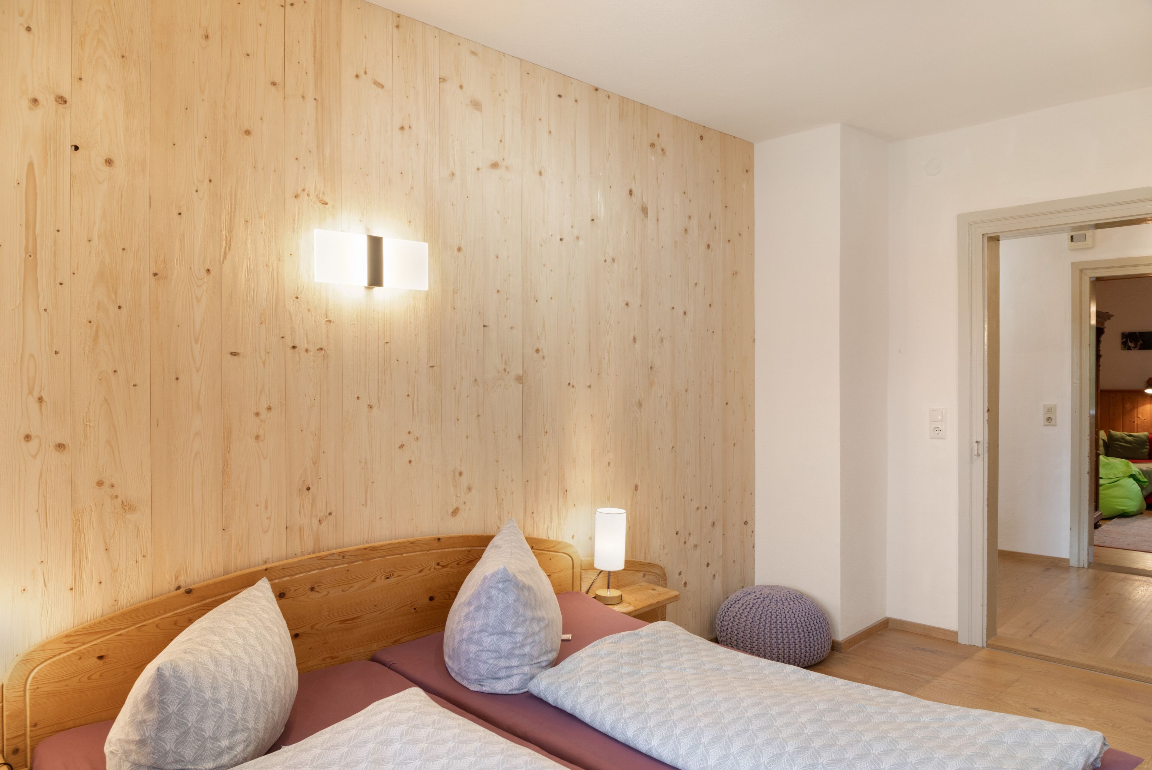 Ein Schlafzimmer mit hölzernem Kopfteil, einem Bett mit gemusterten Betttextilien, einem Nachttisch mit Lampe sowie einer Wandleuchte.