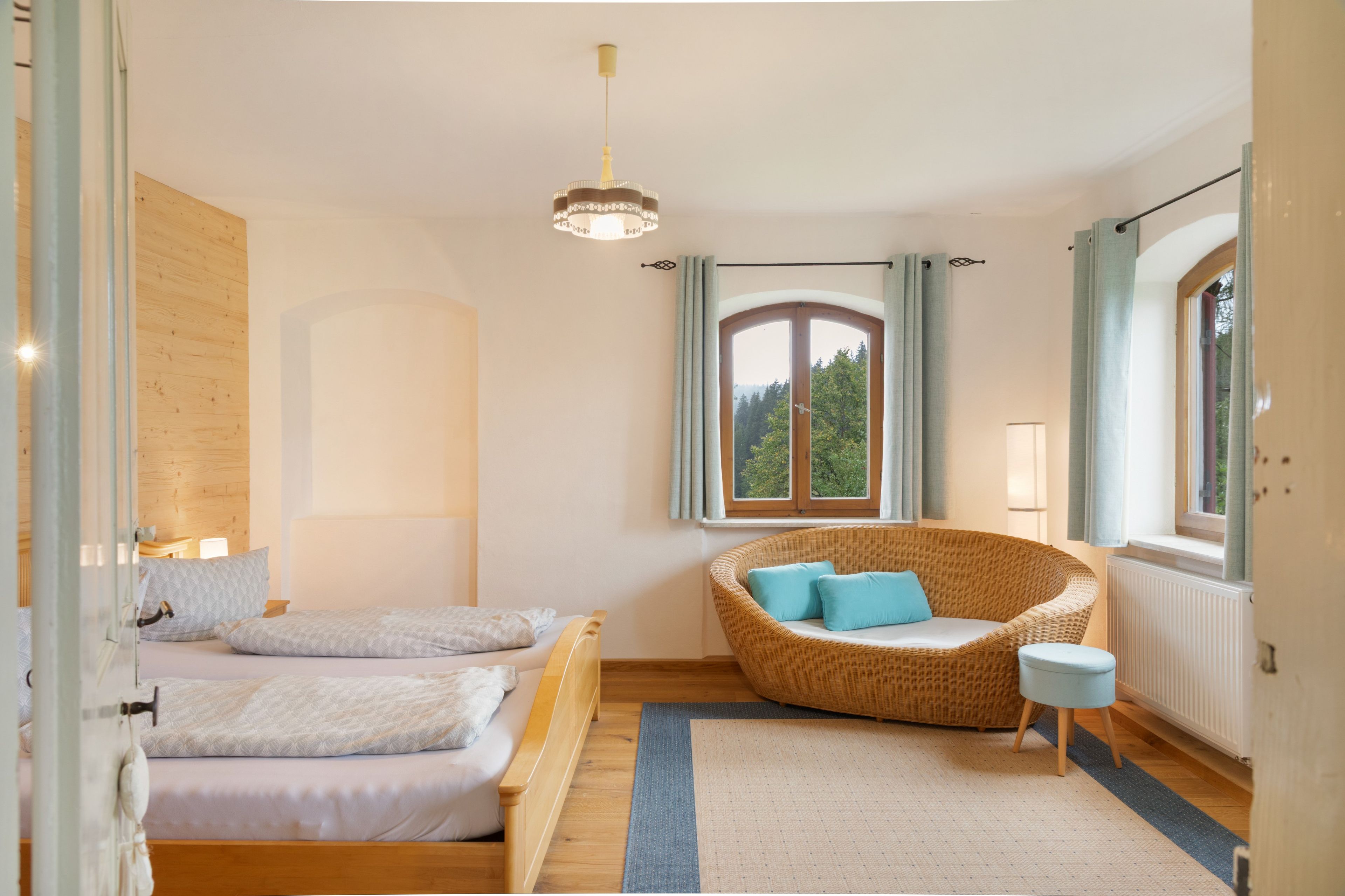 Ein Schlafzimmer mit einem Bett, einem Korbstuhl, einem Fenster mit Vorhängen und einer Pendelleuchte.