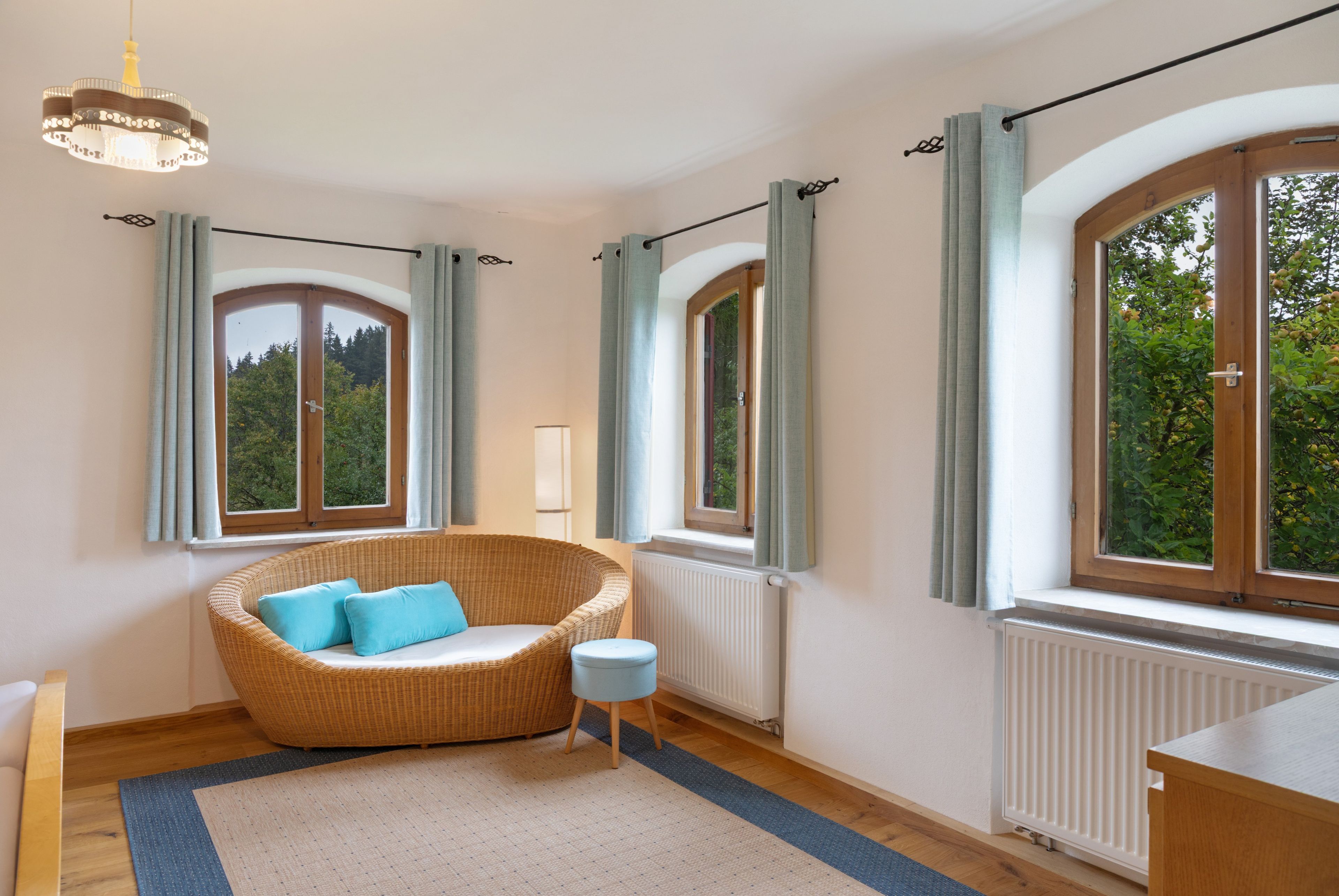 A room with a large arched window, a wicker chair with cushions, a small stool, and a radiator.