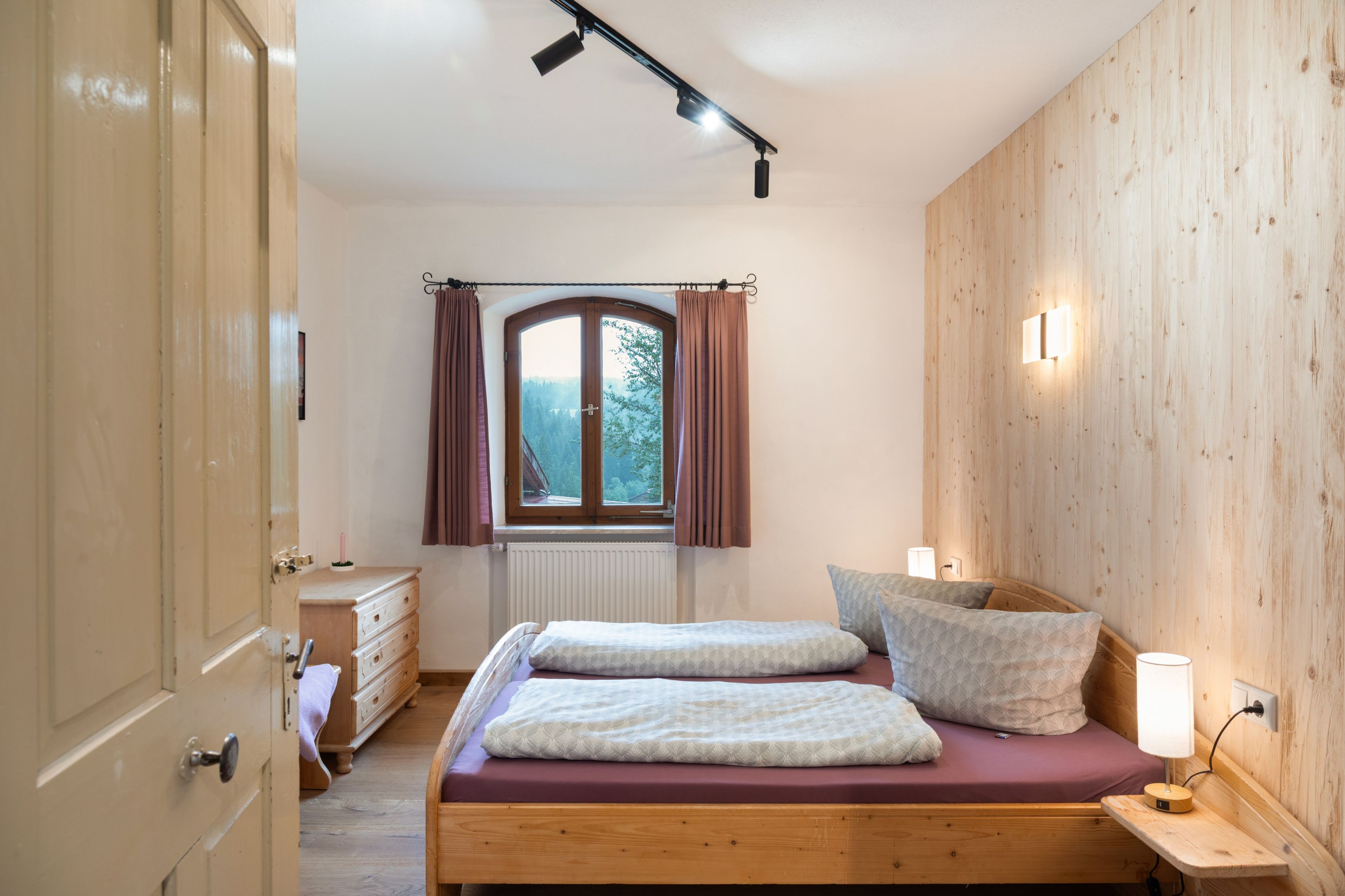 A bedroom with a wooden bed, a window with curtains, a bedside table with a lamp, and a radiator.