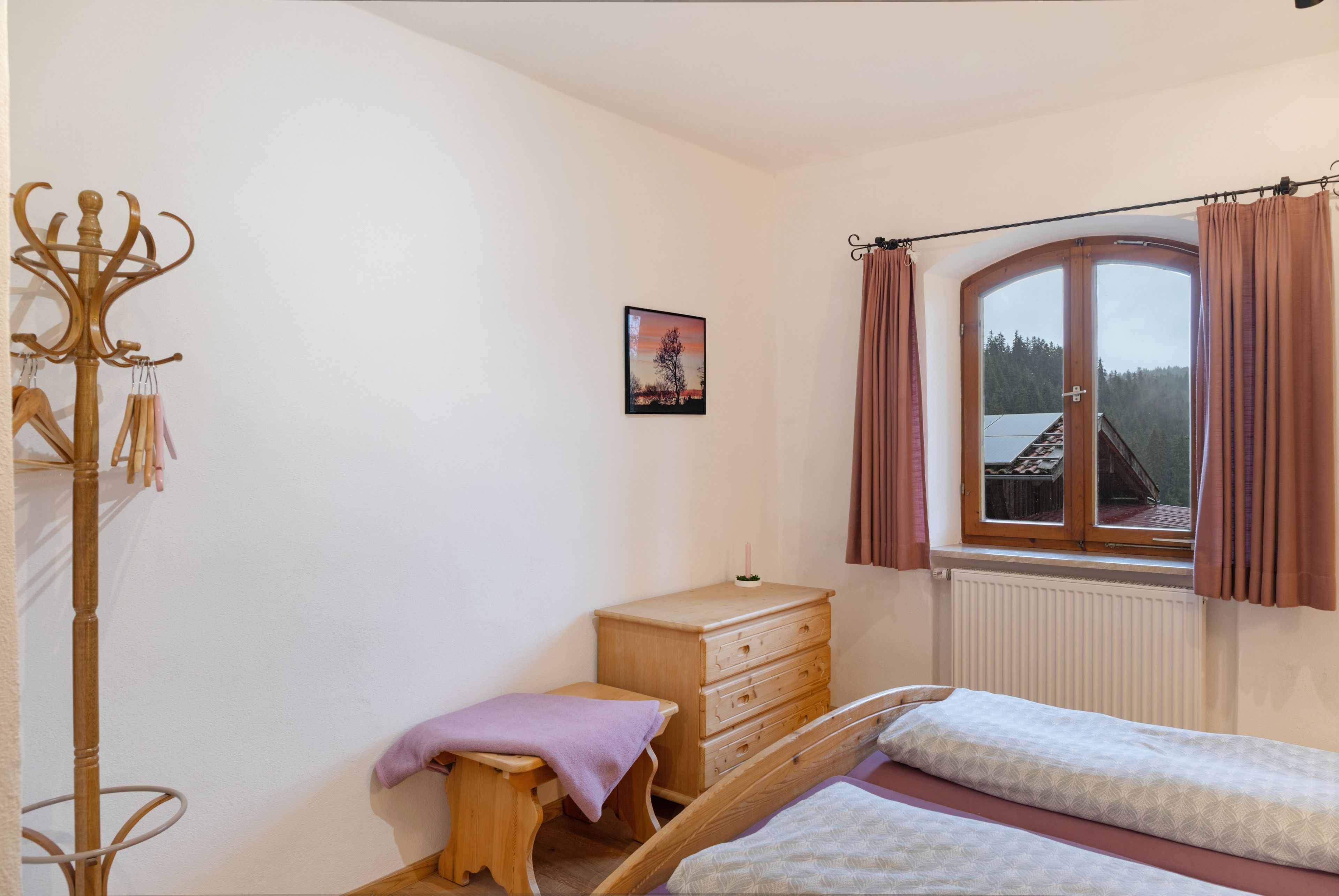 Une chambre à coucher avec un lit en bois, une commode, un petit tabouret, un porte-manteau et une fenêtre avec des rideaux.