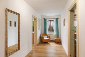 Un passadís amb un mirall gran, una cadira de vimet, una taula petita i una finestra amb cortines.