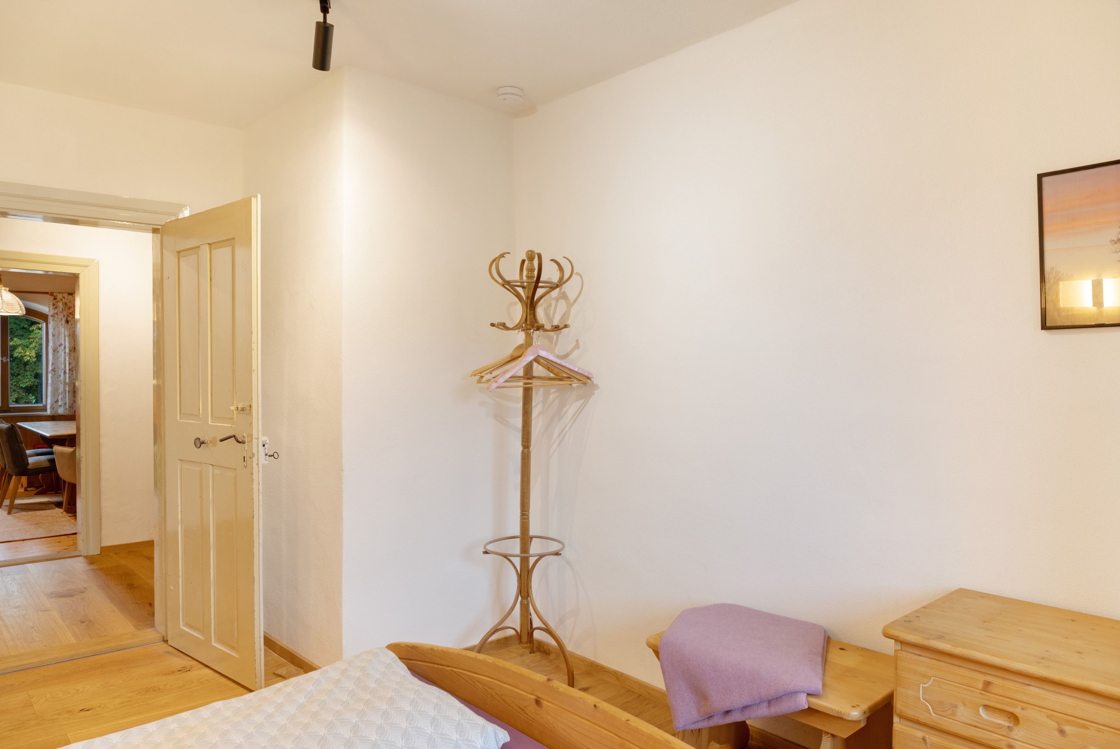 Quarto com cama, cómoda de madeira, cabideiro e porta que dá para outro espaço.