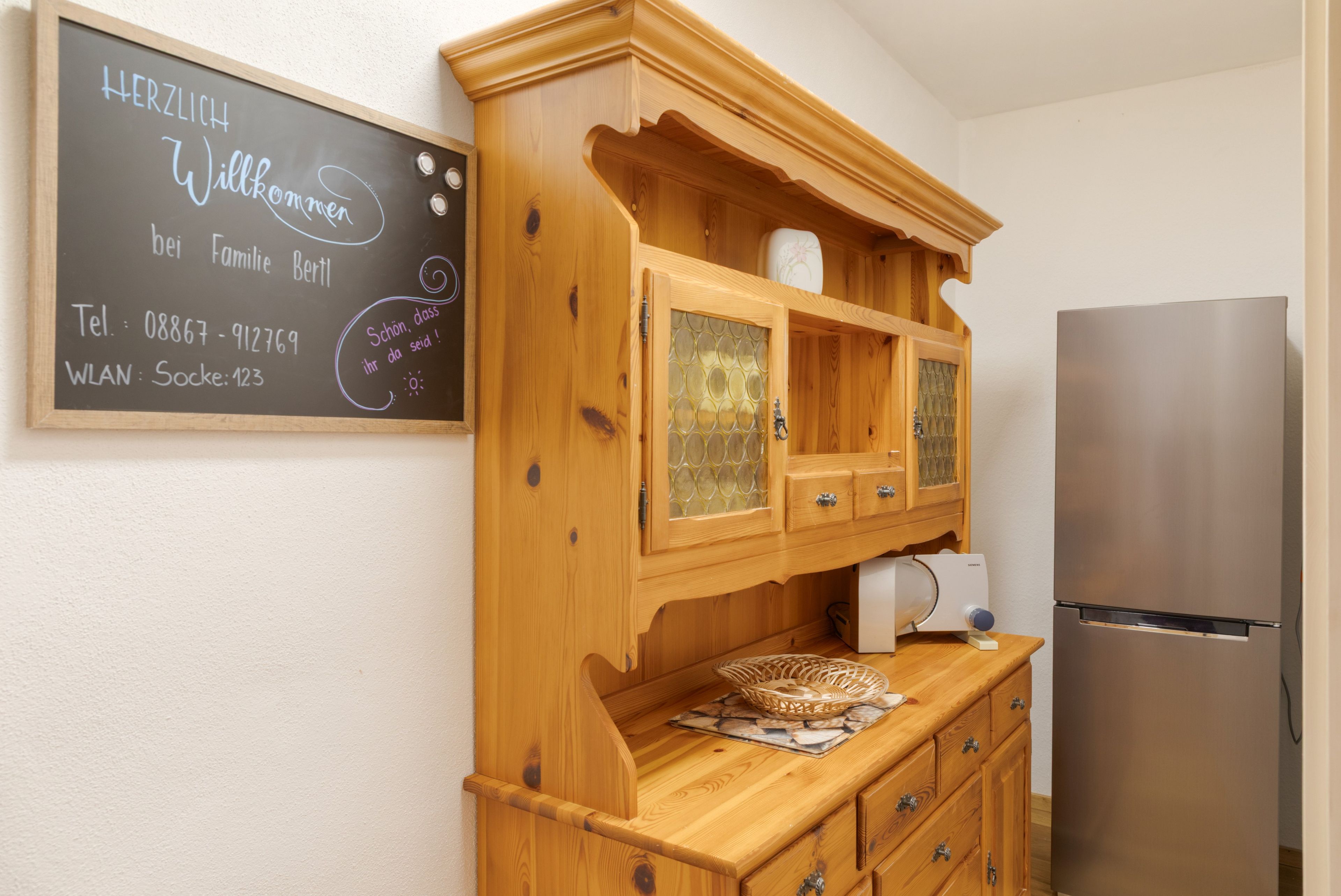Cozinha com armários de madeira, geladeira e quadro negro com informações de contato.