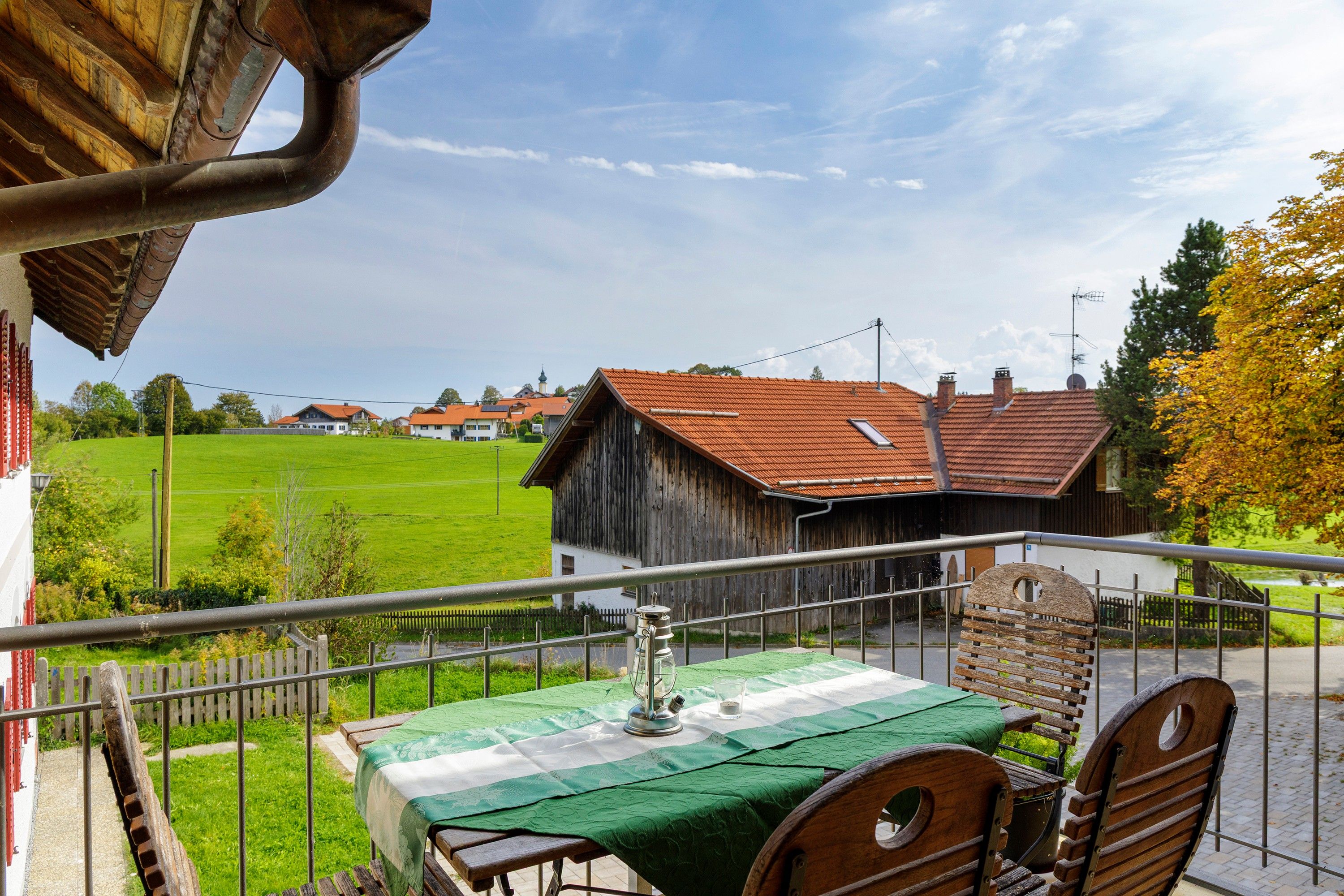 A balcony with a table set for two, overlooking a rural landscape with houses and a green field.
