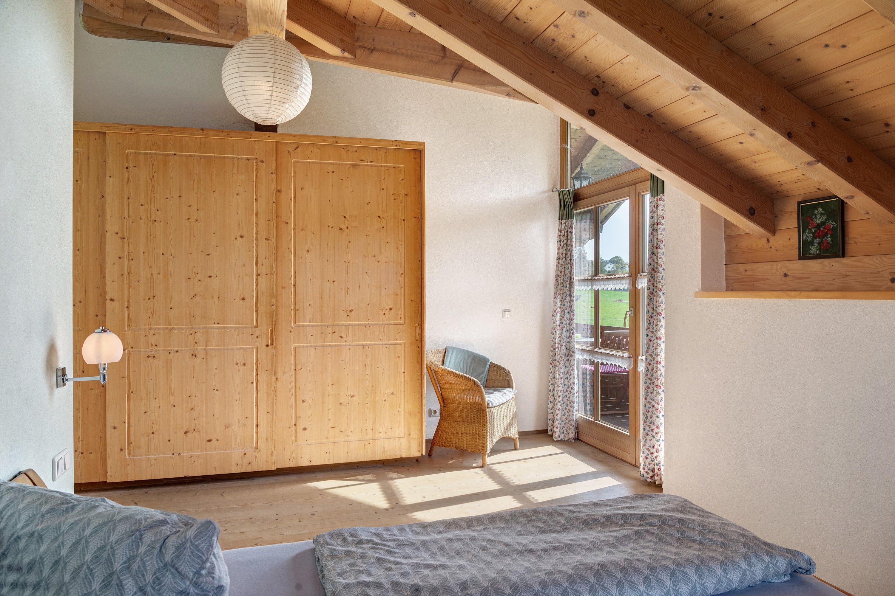 Ein Zimmer mit Holzboden, einer großen Holztür, einem Korbstuhl und einem Bett mit einem gemusterten Überwurf.