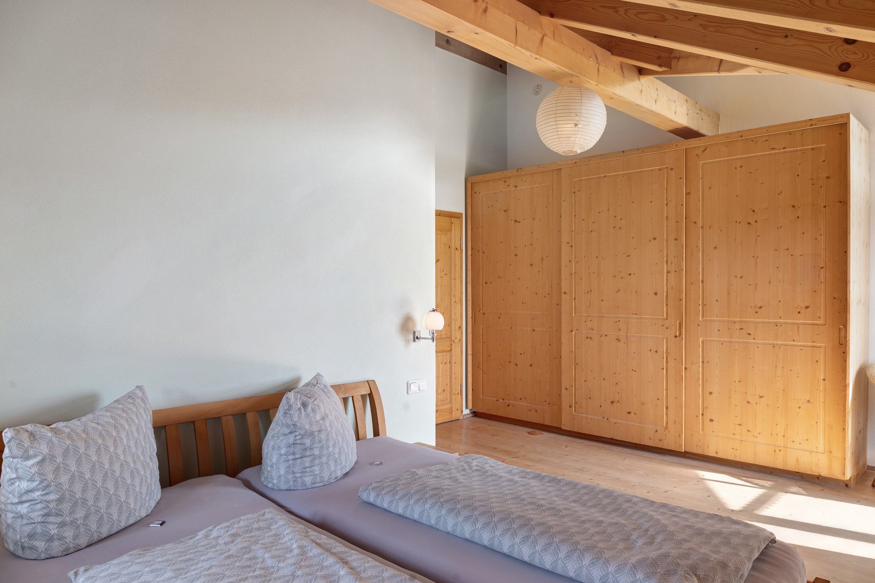 Un dormitorio con cama, dos almohadas, un armario de madera y una lámpara fijada a la pared.