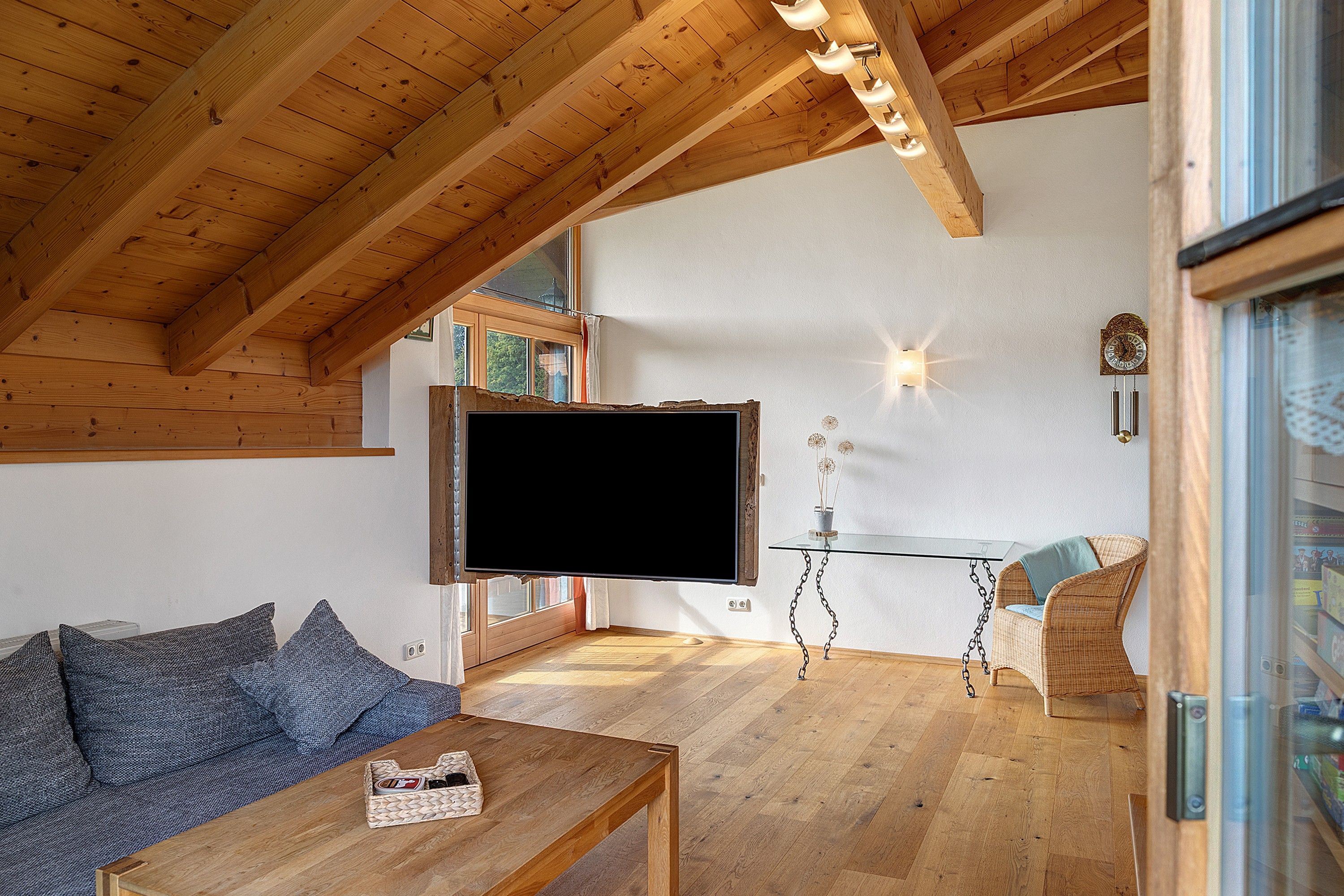 Una sala de estar acogedora con techo de madera, un televisor de pantalla plana montado en la pared, una mesa de centro de vidrio y un sofá de mimbre.