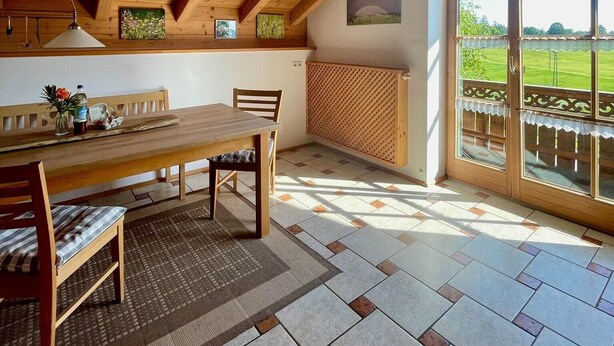 Un comedor con una mesa y sillas de madera, un banco, un radiador y vistas a través de un ventanal.