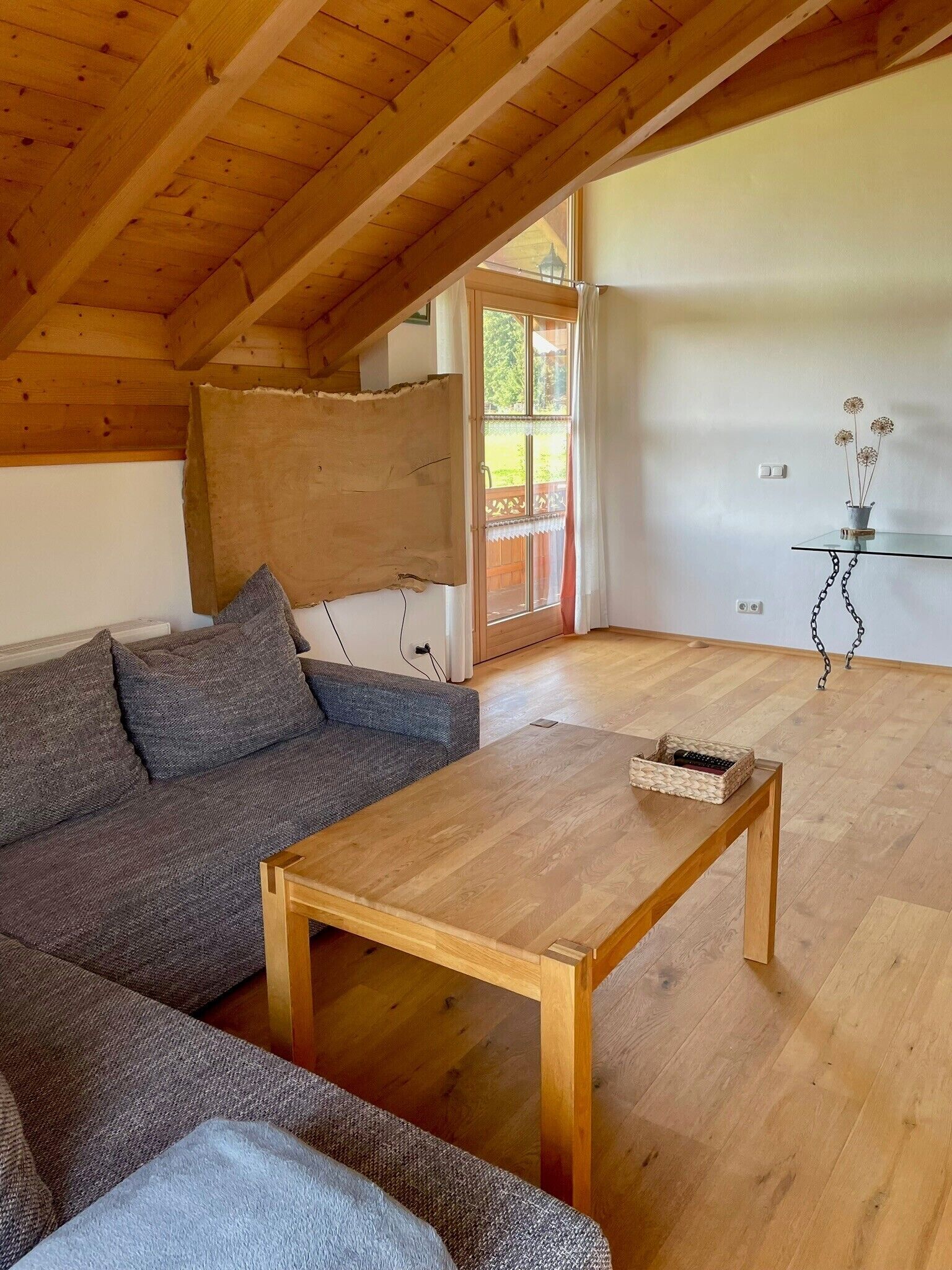 Ein gemütliches Wohnzimmer mit Holzbalkendecke, einer Couch, einem Couchtisch und Blick durch eine Glastür.