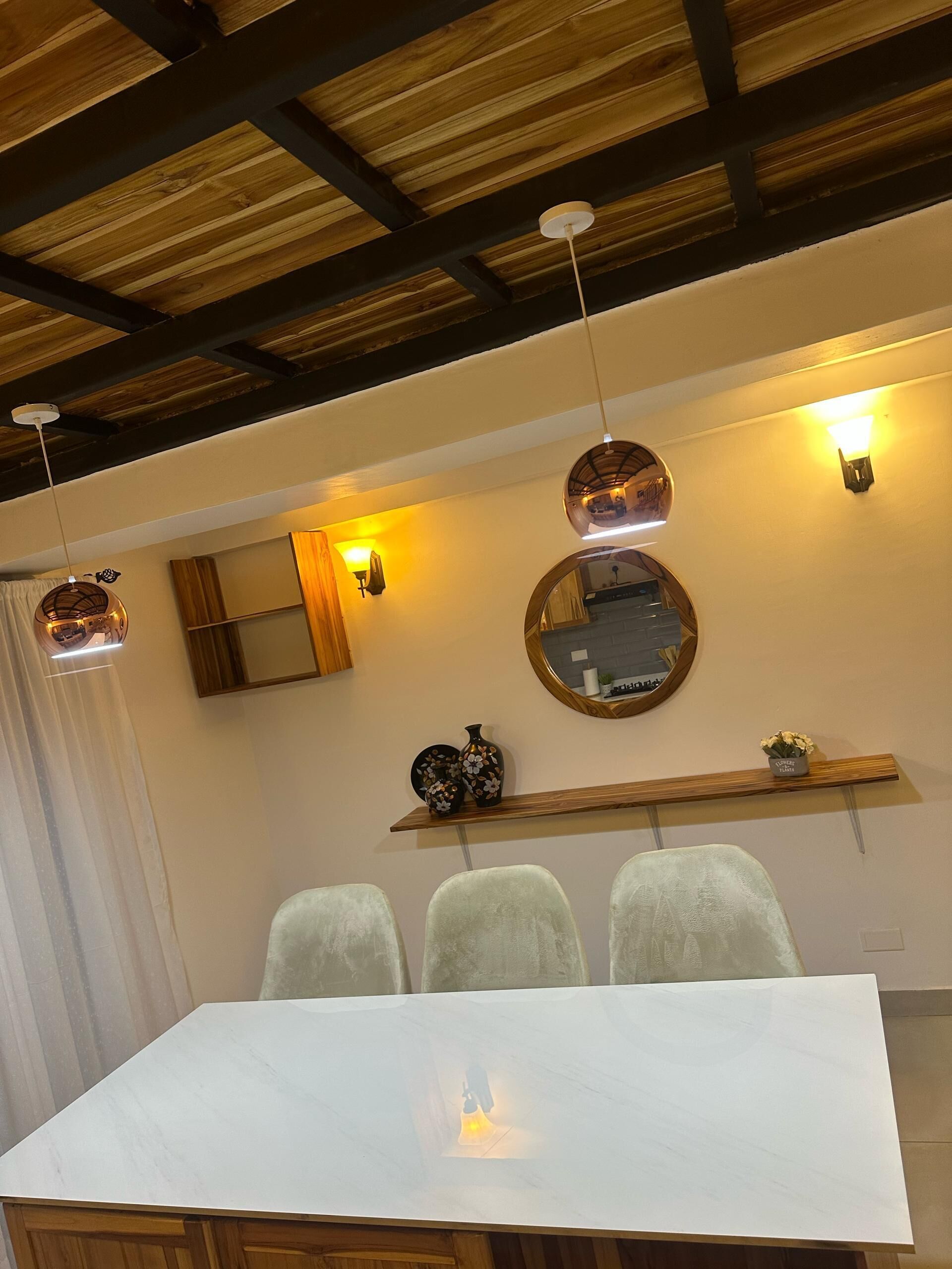 A dining area with a white table, three chairs, a wooden ceiling, and wall-mounted light fixtures.