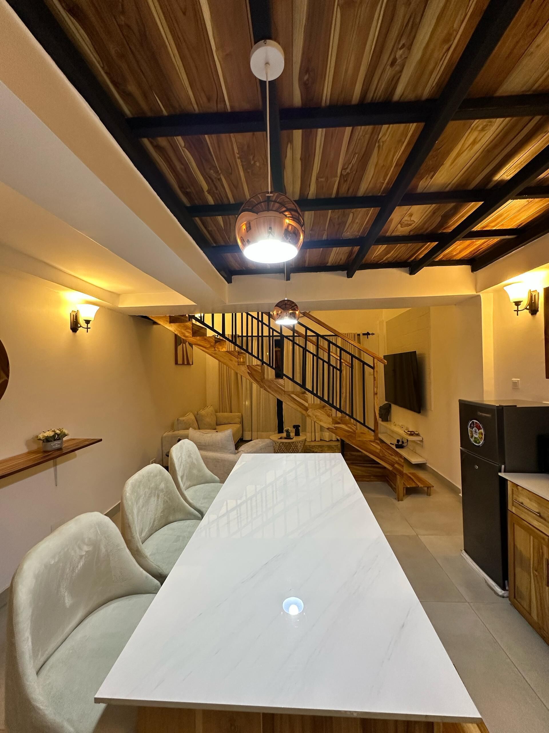 A dining area with a long table and chairs, a staircase with wooden railings, and a kitchen area with a refrigerator.