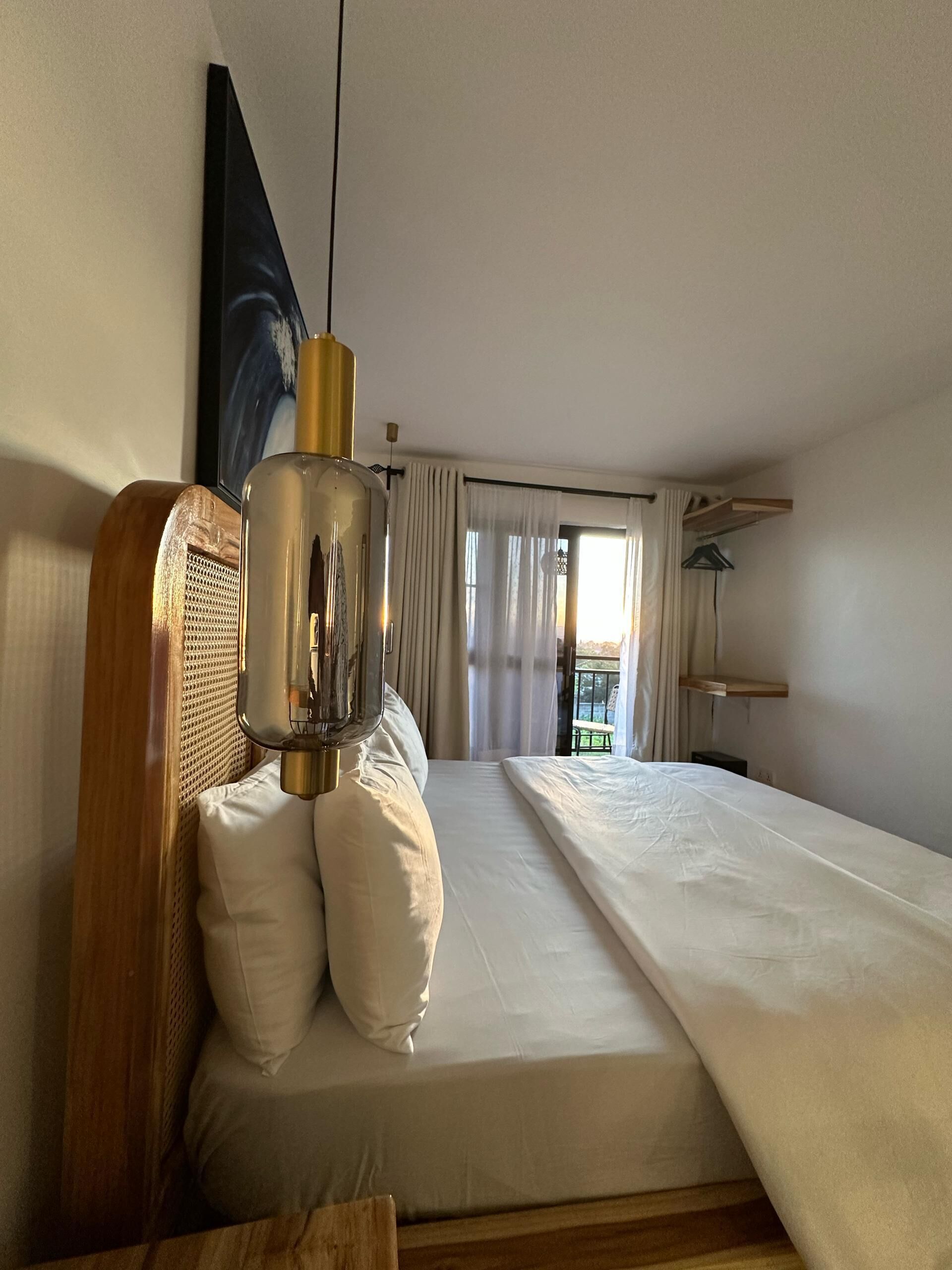 A hotel room with a bed, a hanging light fixture, a window with curtains, and a view of the outdoors.