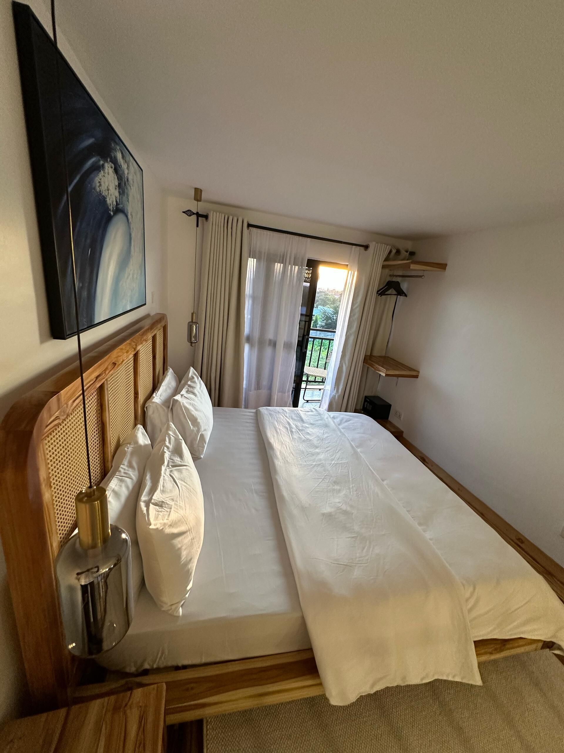 A hotel room with a bed, a painting, a lamp, and a view of the outdoors.
