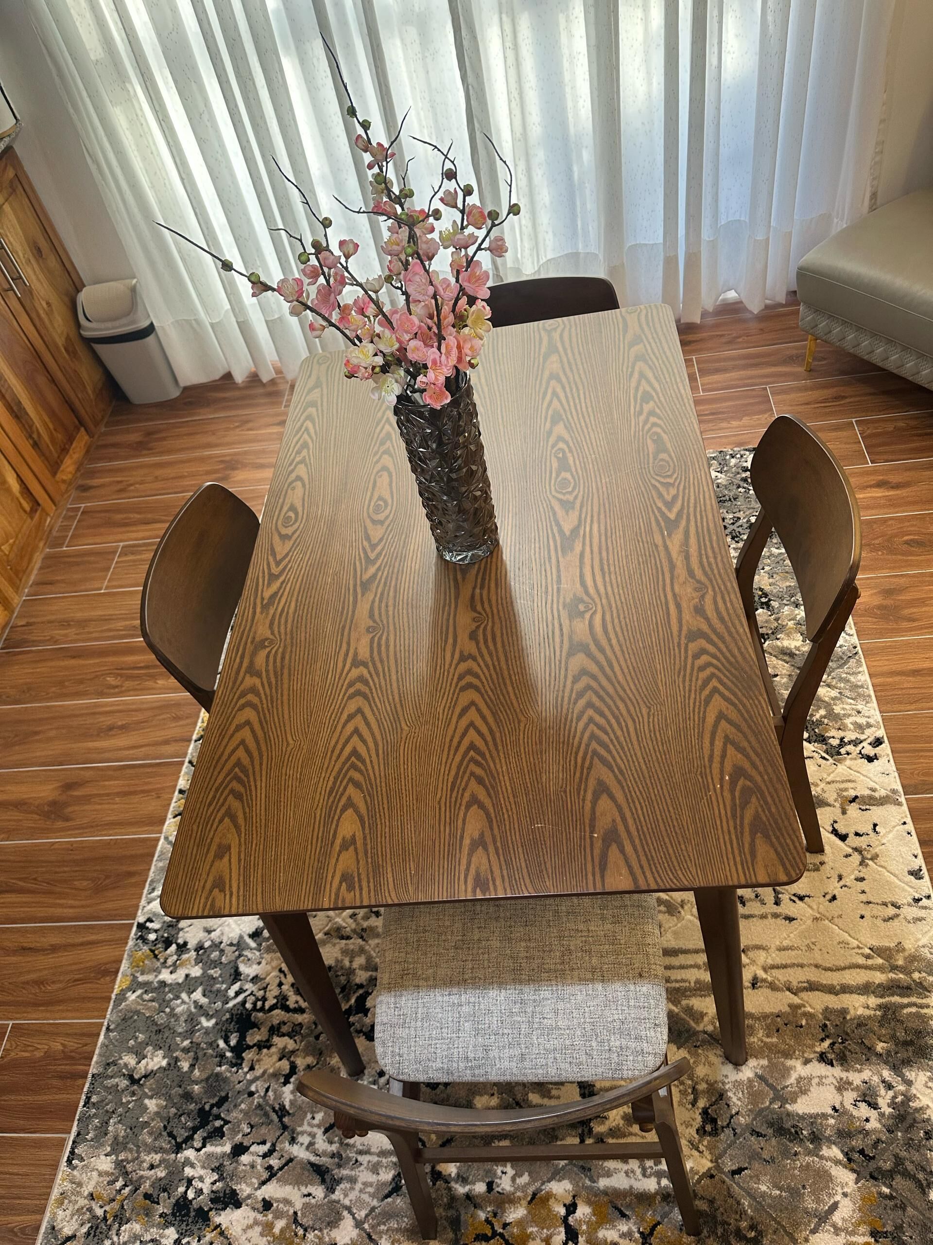 A dining table with a vase of flowers, surrounded by chairs and a patterned rug.