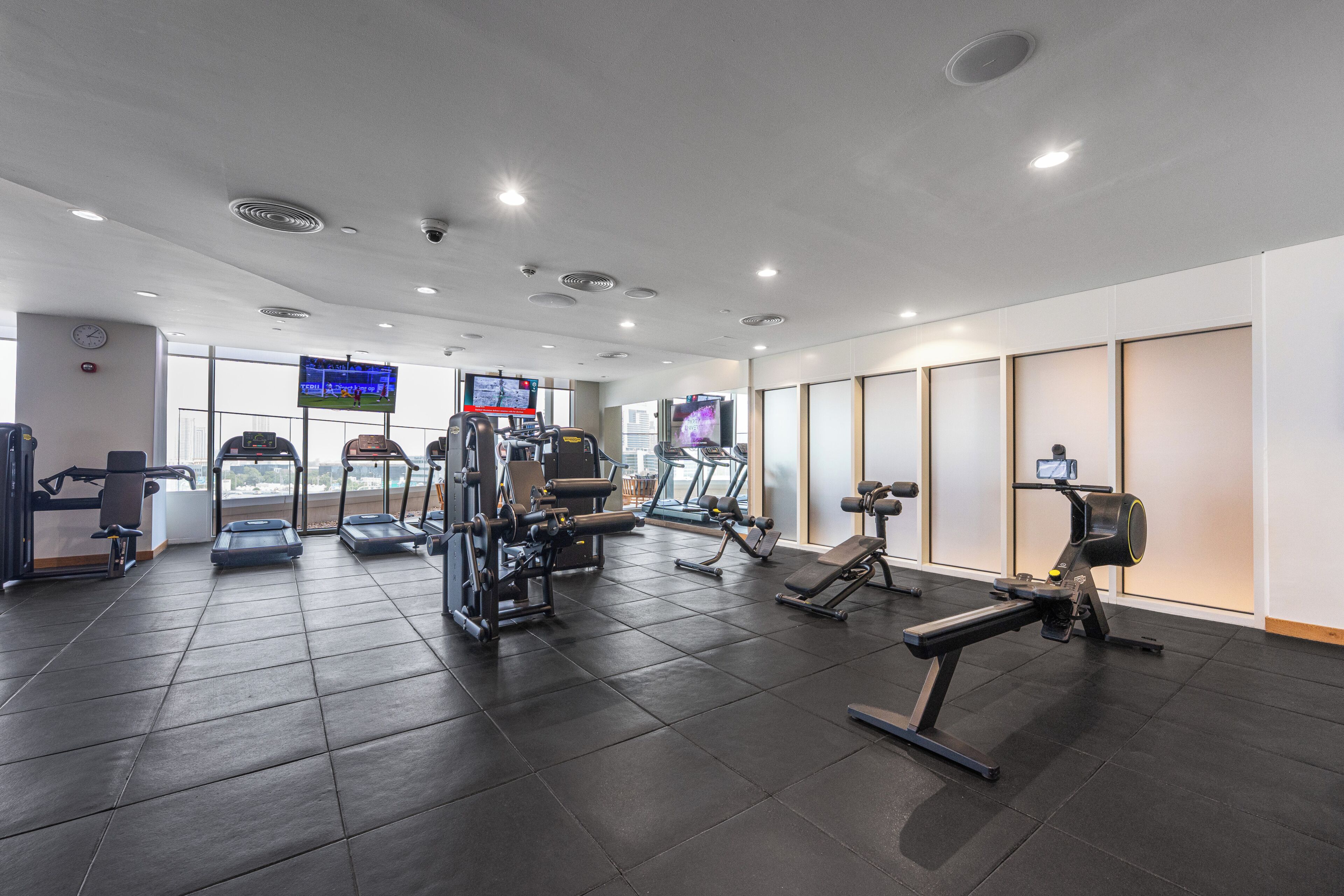 Ett gym med olika typer av träningsutrustning, inklusive löpband och styrketräningsmaskiner, och en stor skärm som visar en sportevenemang.