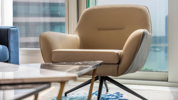A modern chair with a high backrest and metal legs, positioned near a window with a view of a cityscape. - StayShort - Dubai Media City sleeps 2 with Amazing Sea Views! (Media City)