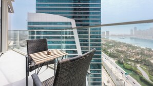 A balcony with a wicker chair and table, overlooking a cityscape with modern buildings and a river. - StayShort - Dubai Media City sleeps 2 with Amazing Sea Views! (Media City)