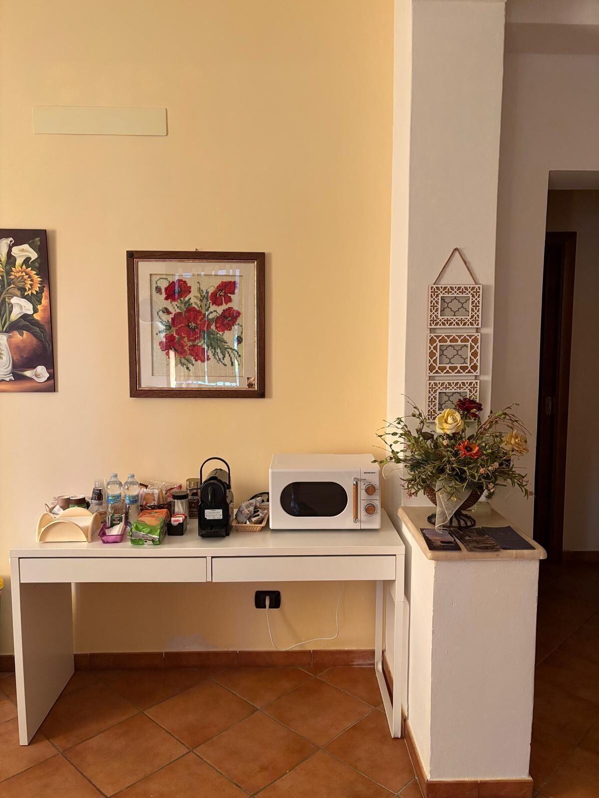 A room with a white desk, a microwave, a basket of fruits, a framed picture of flowers, and a small hanging decoration.