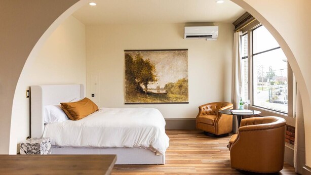 Une chambre d’hôtel avec un lit, un tableau, une chaise, une table et une fenêtre donnant sur un paysage.