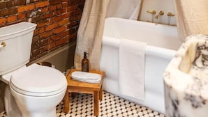 Une salle de bain avec un mur en briques, des toilettes blanches, une baignoire avec une pomme de douche dorée et un petit tabouret en bois.