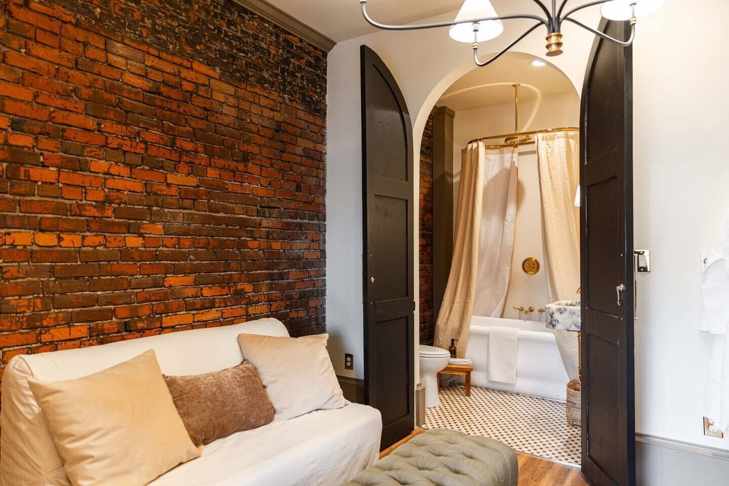 A room with a brick wall, a sofa, a small ottoman, and a bathroom with a bathtub, toilet, and shower curtain.