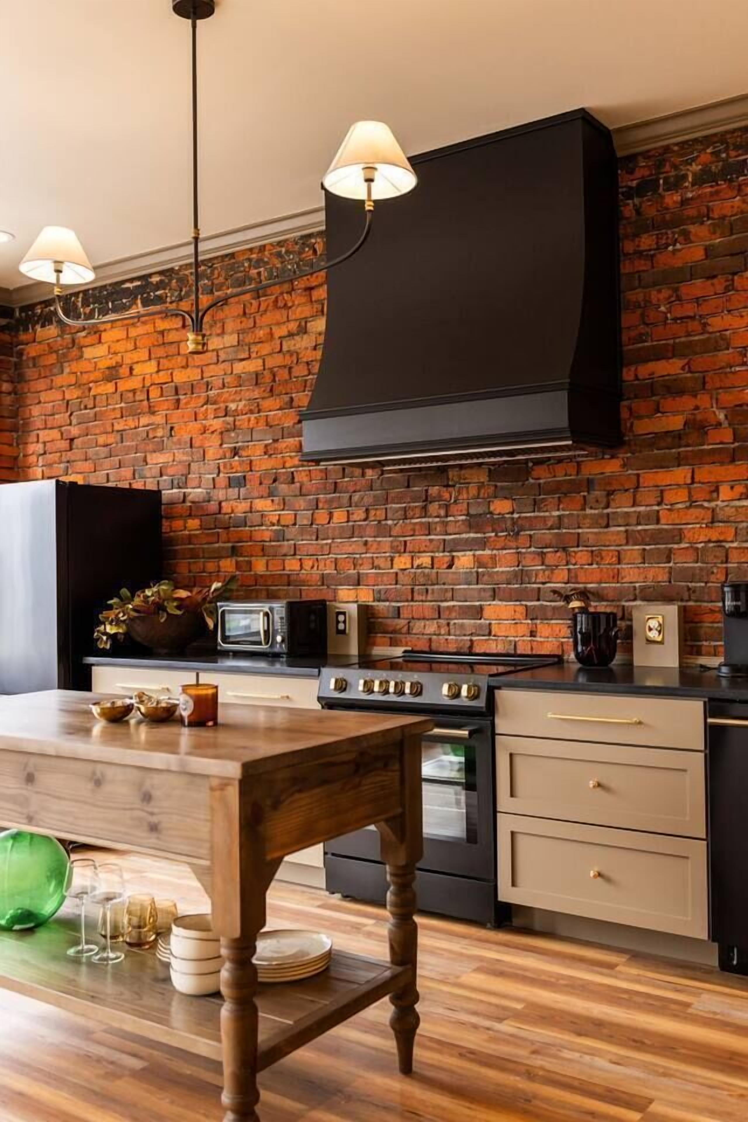 A kitchen with a brick wall, a black range hood, a wooden table, and a hanging light fixture.