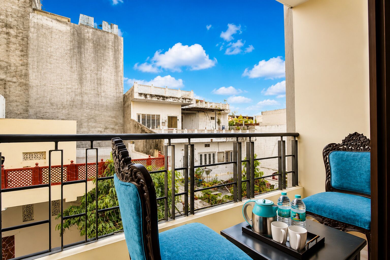 Een balkon met uitzicht op gebouwen, een tafel met een theepot, kopjes en een fles, en twee blauwe stoelen.