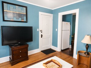 A room with a television on a wooden cabinet, a door, a refrigerator, and a lamp on a side table. - Classic Fargo Retreat Near NDSU & Downtown (Fargo)