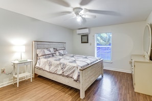 Une chambre à coucher comprenant un lit, une table de chevet avec une lampe, un ventilateur de plafond, un appareil de climatisation, une fenêtre avec des stores et une commode.