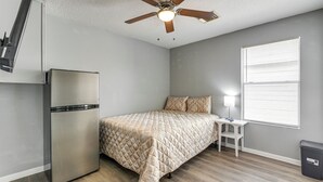 Habitación compacta con cama, una mesita, refrigerador, ventilador de techo y ventana con persianas.