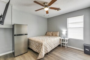 Ein kleines Schlafzimmer mit Bett, Tisch, Kühlschrank, Deckenventilator und Fenster mit Jalousien.