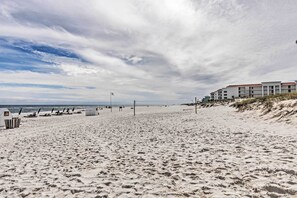 A beach with a row of buildings, a volleyball net, and a lifeguard tower. - Unwind Poolside, Walk to Shore! Orange Beach Condo (Orange Beach)