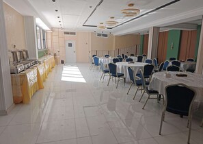 Un salón de banquetes con varias mesas redondas y sillas, una zona de bufé con equipo para servicio de alimentos y un suelo de baldosas.