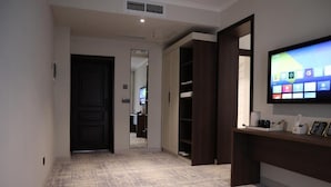 A hotel hallway with a carpeted floor, a dark wooden door, a mirror, a wardrobe, and a flat-screen TV mounted on the wall.