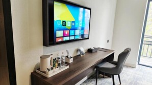 A hotel reception area with a flat-screen TV mounted on the wall displaying a colorful interface, a wooden reception desk with a chair, and a small coffee station.