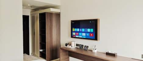 Habitación de hotel con cama, escritorio con televisor de pantalla plana, armario de madera y mesita de noche con lámpara y teléfono.