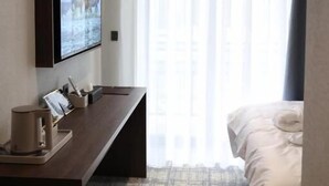 A hotel room with a bed, a desk with a computer, a TV mounted on the wall, and a window with sheer curtains.