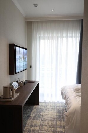 A hotel room with a bed, a desk with a computer, a TV mounted on the wall, and a window with sheer curtains.