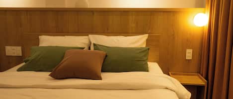 A hotel room with a wooden headboard, a bed with white bedding and green and brown pillows, a bedside table with a lamp, and a wall socket.