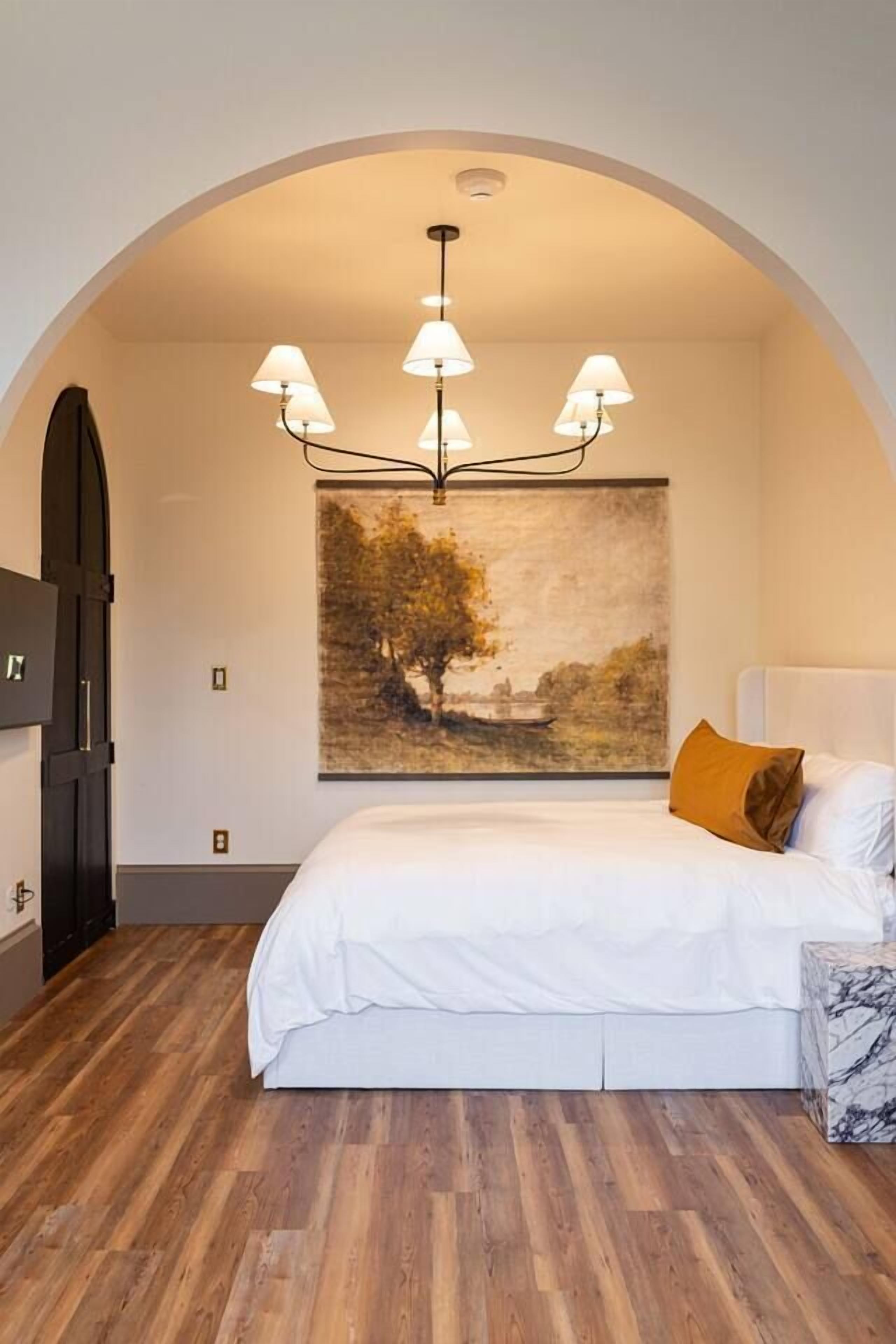 A bedroom with a wooden floor, a bed with white bedding, a painting on the wall, and a chandelier.