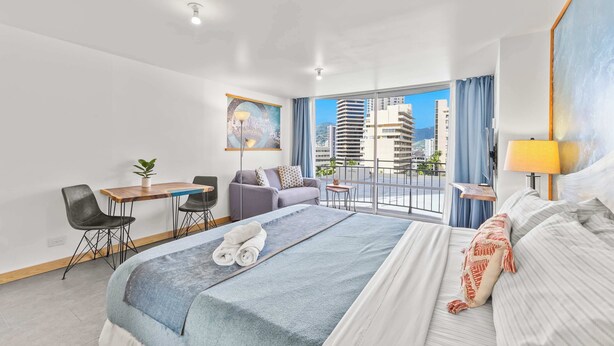 A hotel room with a bed, a desk, a chair, a small table, a potted plant, and a view of buildings through the window. - Modern Designer Studio in Waikiki near Ala Moana (Honolulu)